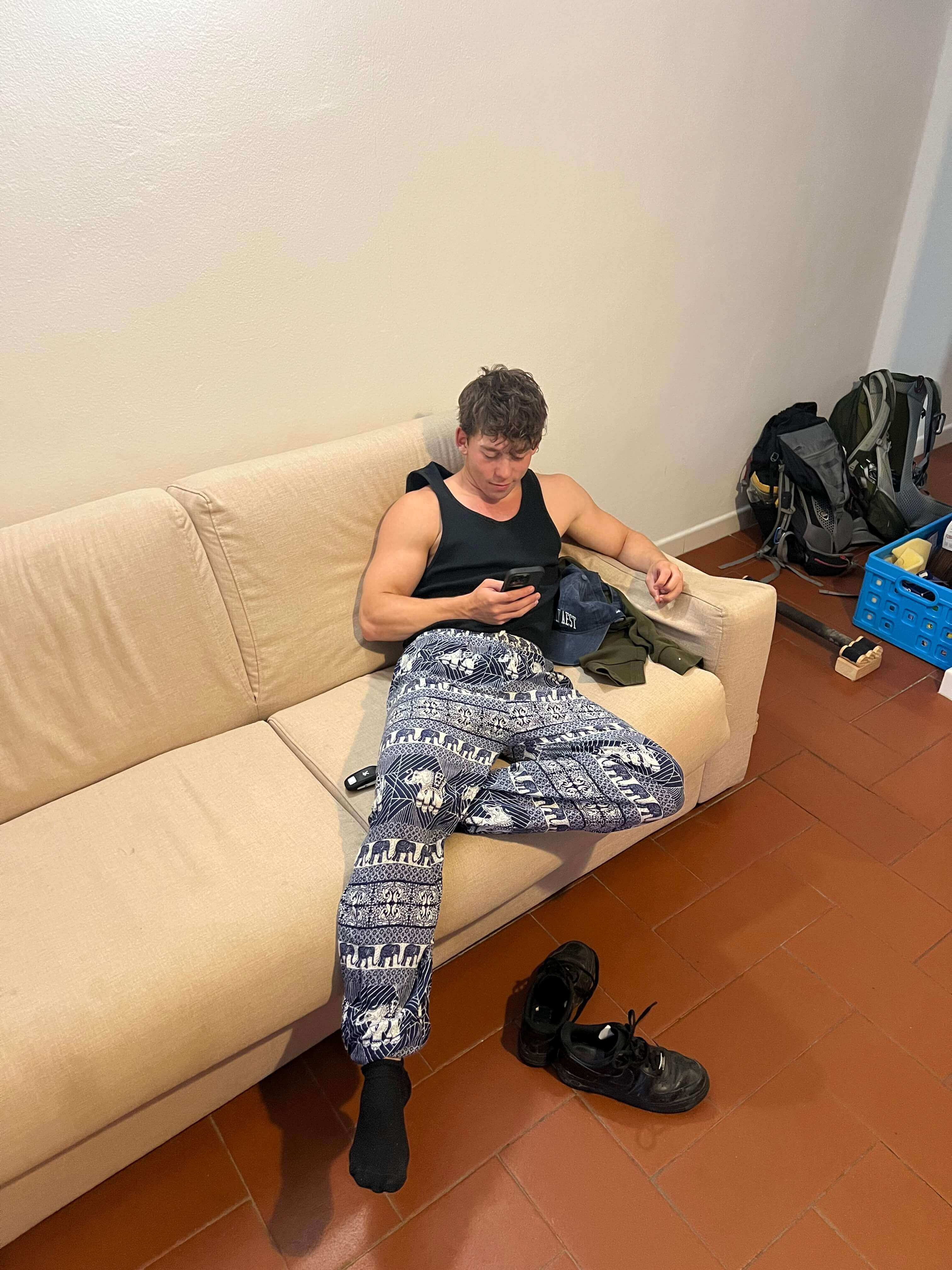 Me chilling on the couch with my mobile in my hand, wearing a black tank top and blue elephant trousers.